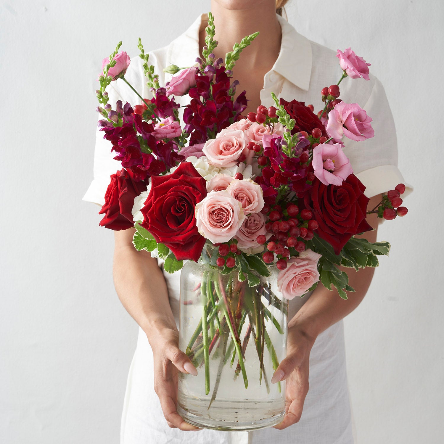 Woman wearing all white holding large red, pink, and red arrangement including roses, lisianthus and snapdragons.