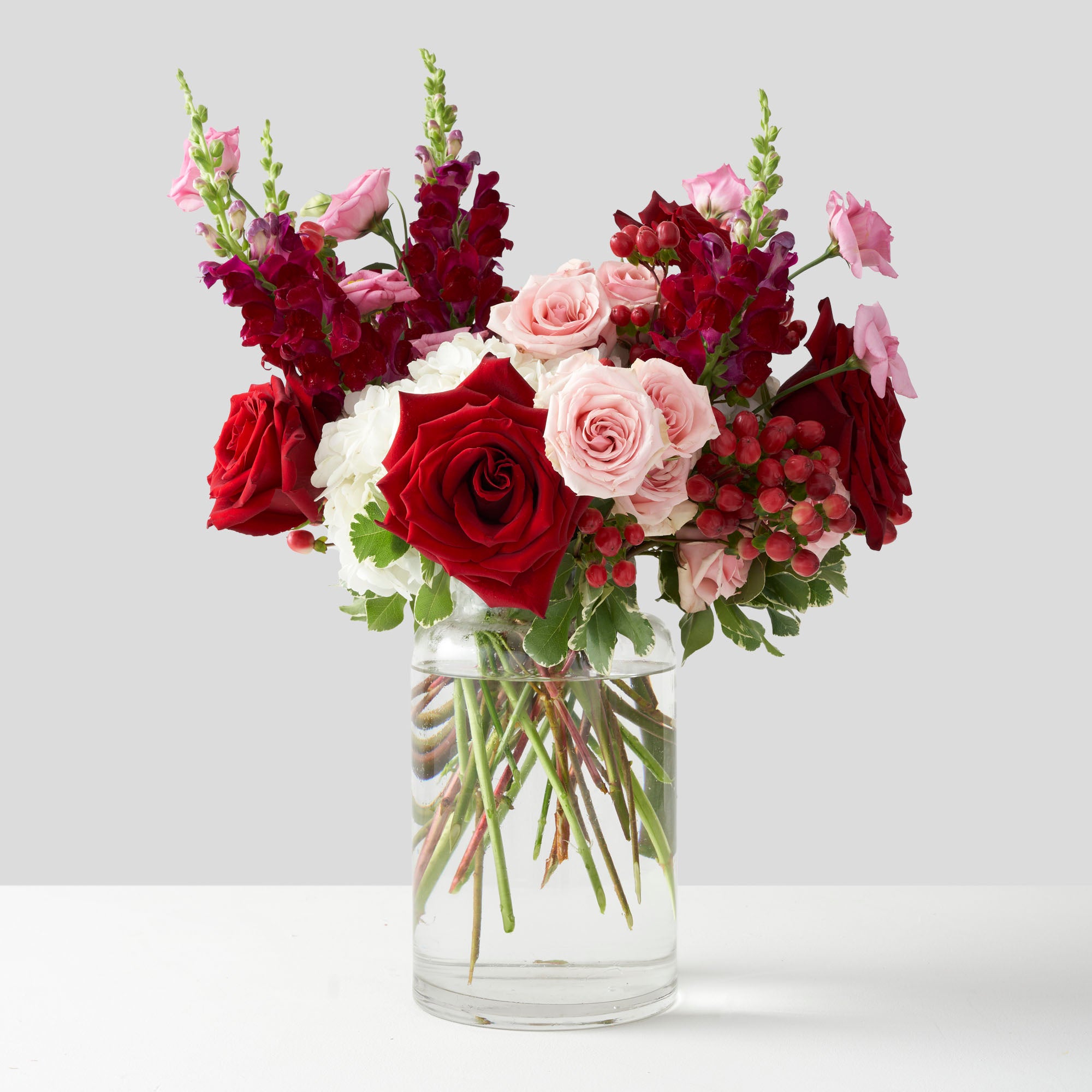 Red roses, pink roses, pink lisianthus and red snapdragons arranged in tall clear glass vase on white background.