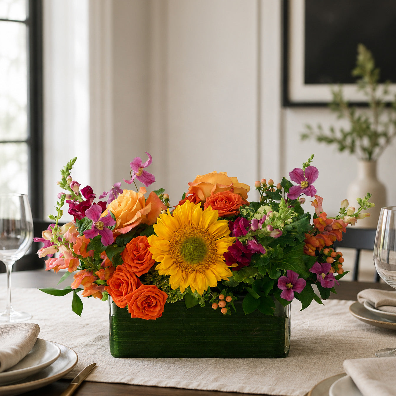 A vibrant floral centerpiece in a rectangular leaf lined vase, featuring a large yellow sunflower, orange roses, and mauve clematis, hot pink snapdragons, styled on a dining table with place settings and wine glasses.