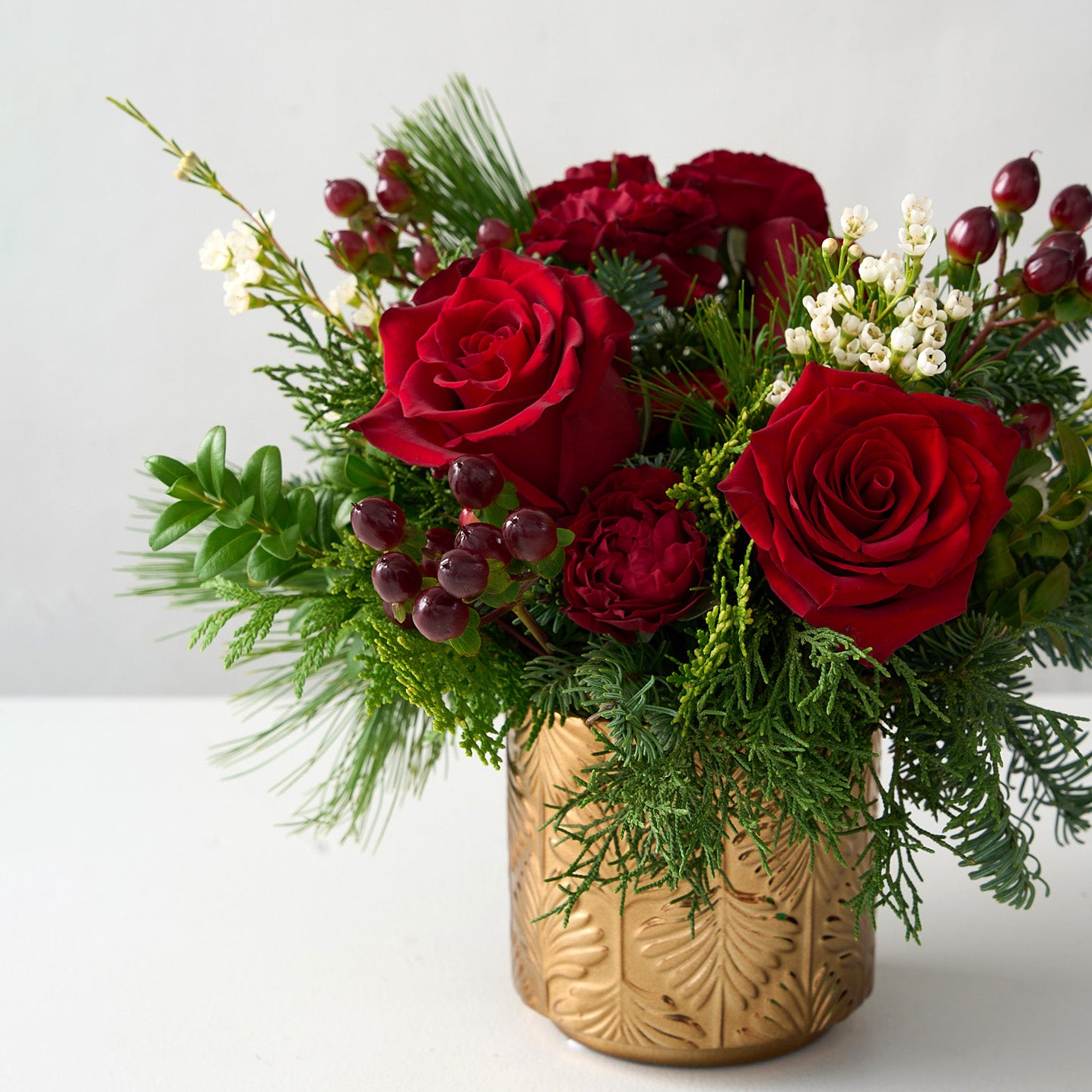 off center picture of floral arrangement with red roses, hypericum, carnations and fresh boxwood, cedar branches with wax flowers in a gold pot