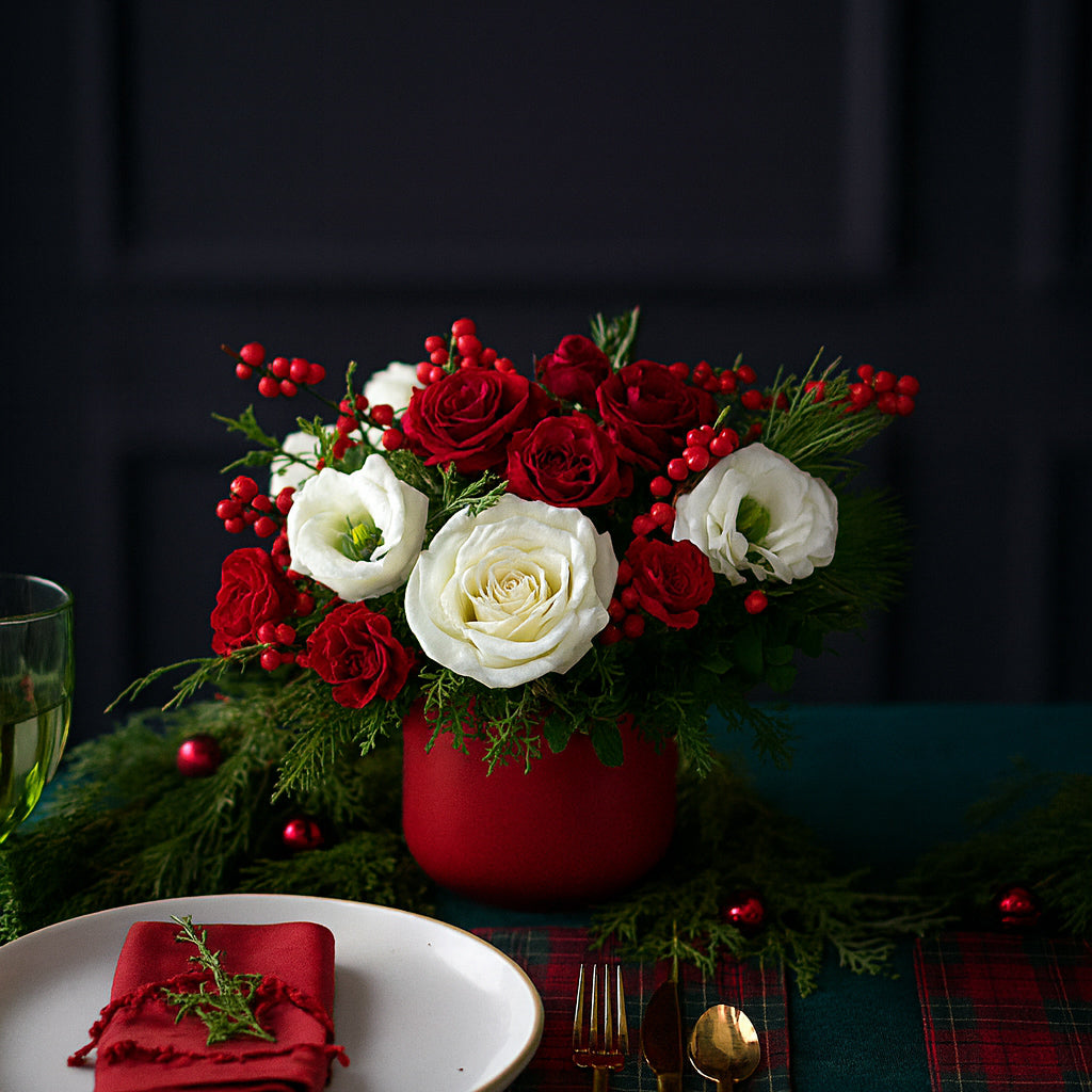 Small christmas arrangement with red and white roses, hypericum berries, winter greens in red ceramic pot on dinner table with holiday decorations