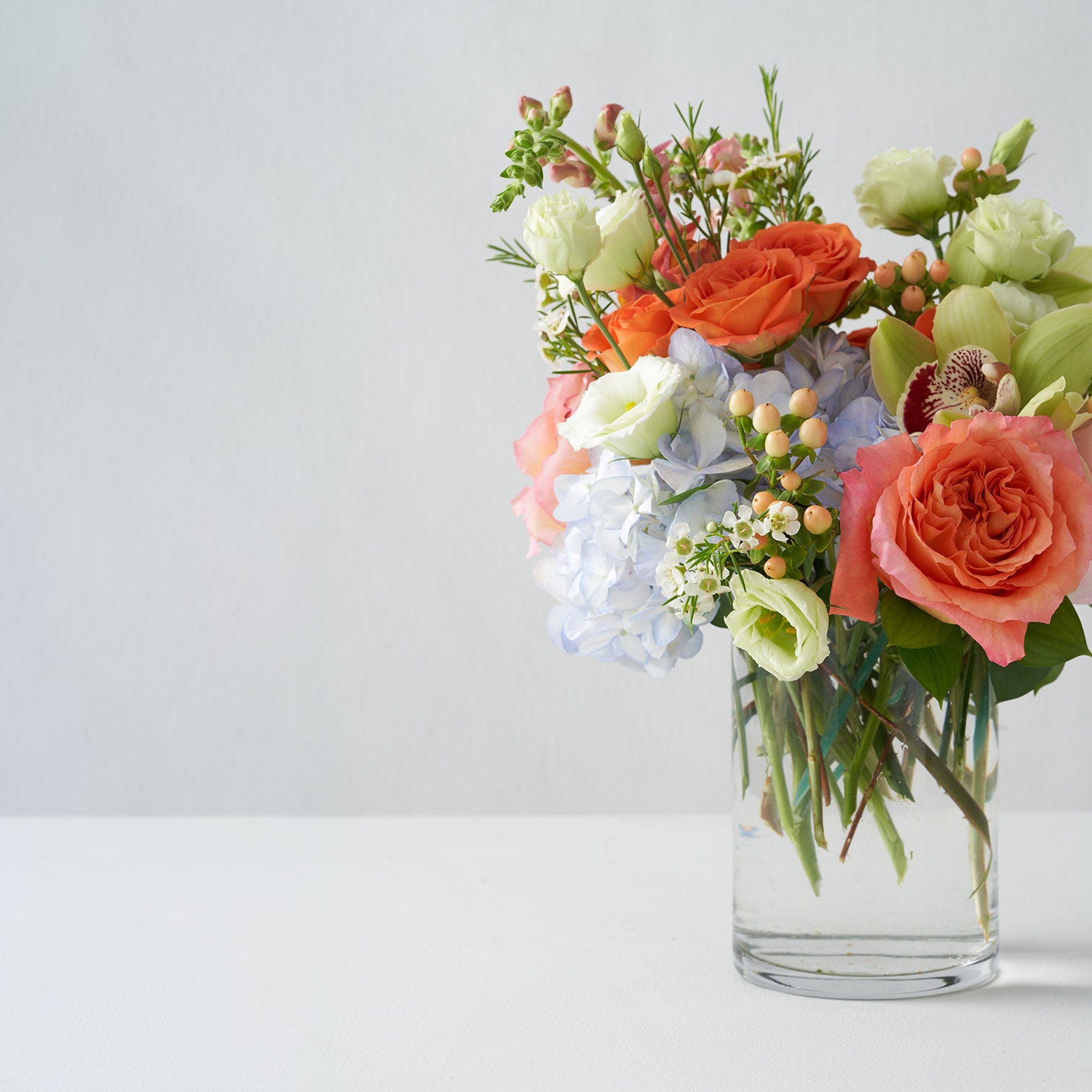 Glass vase of orange roses, peach hypericum berries, blue hydrangea and green cymbidium orchids on white background