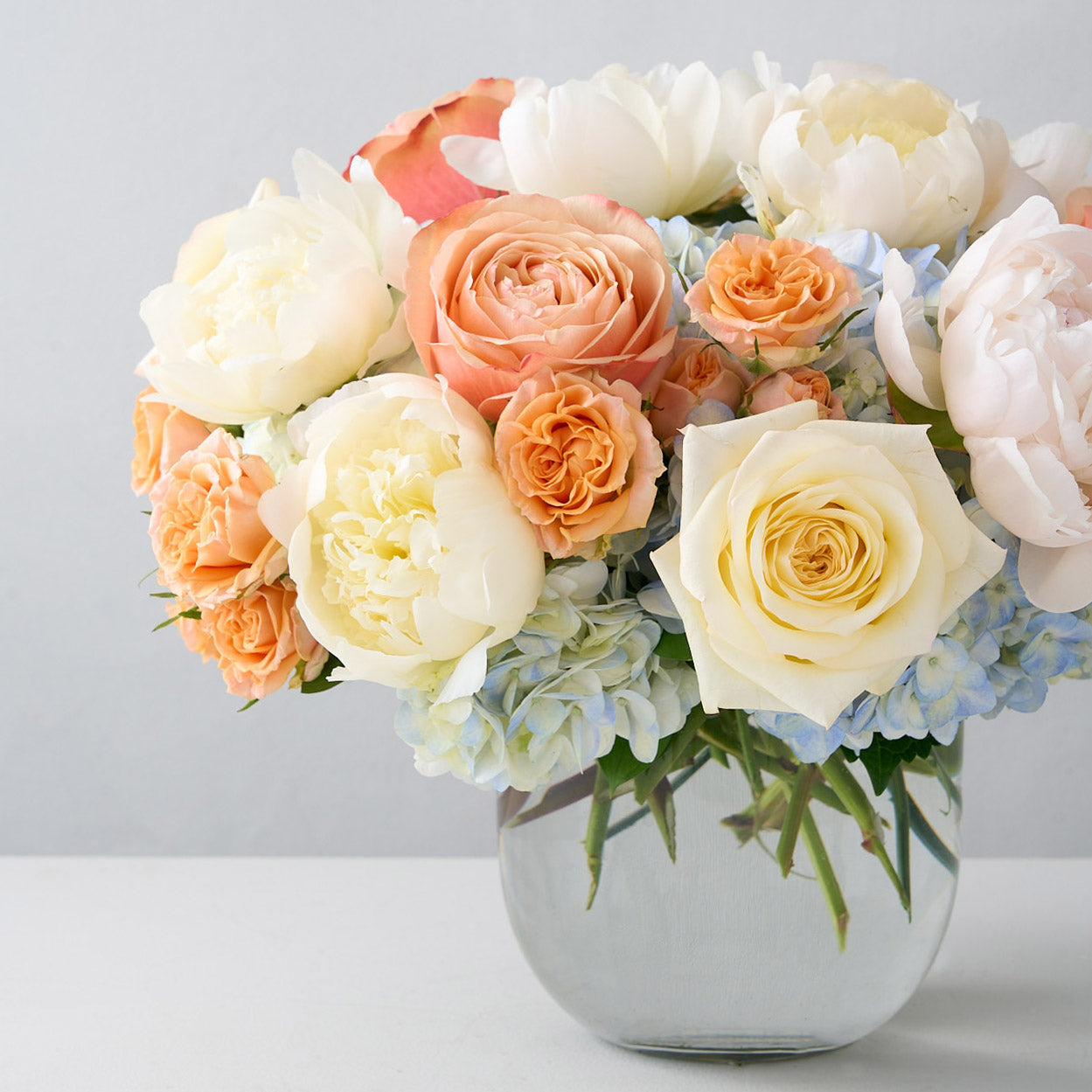 Glass vase filled with a bouquet of white and peach roses, cream peonies, and light blue hydrangeas.