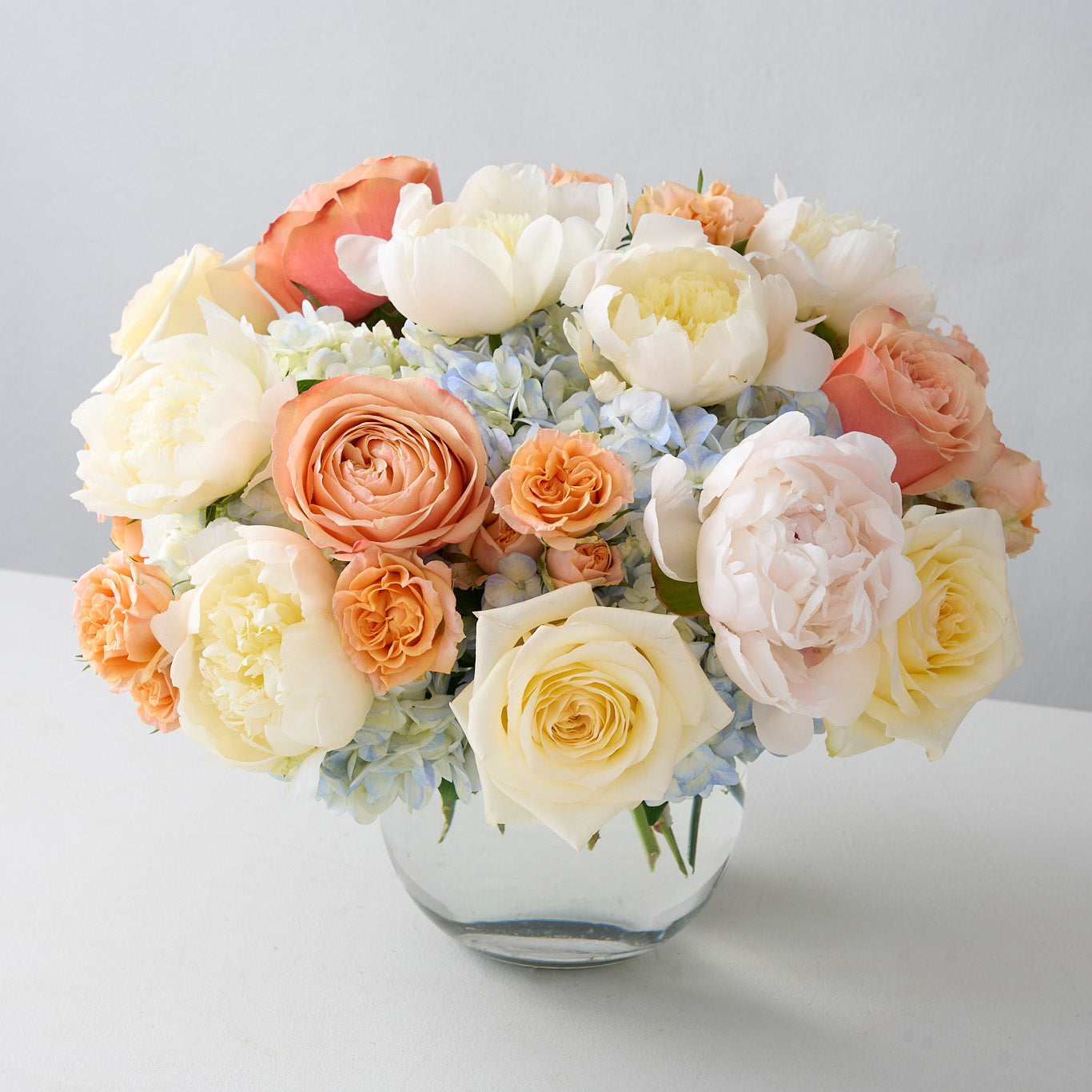 A glass vase filled with a bouquet of white and peach roses, cream peonies, and light blue hydrangeas.