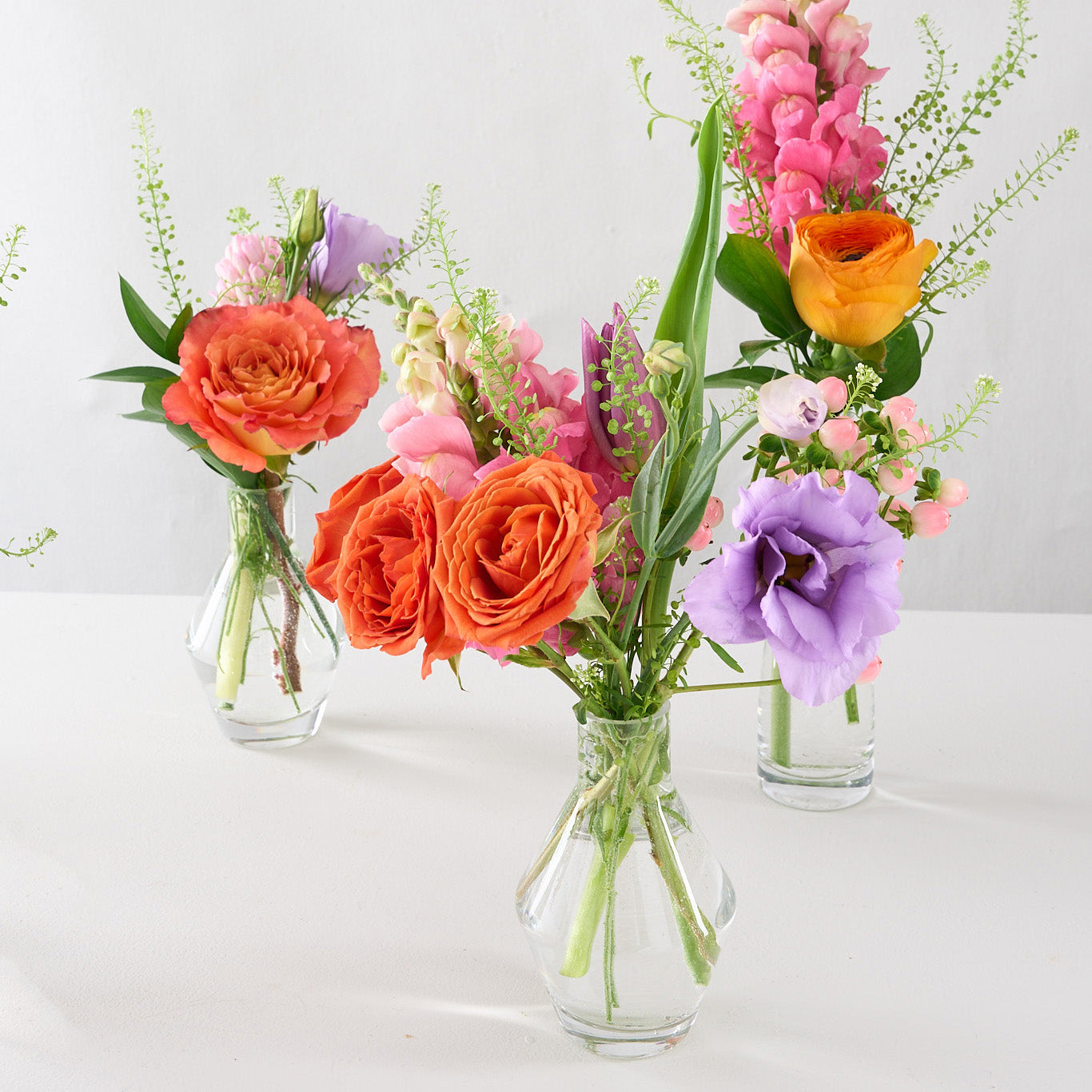 A series of five glass bud vases, each containing an arrangement of colorful spring flowers, arranged in a line on a light background.