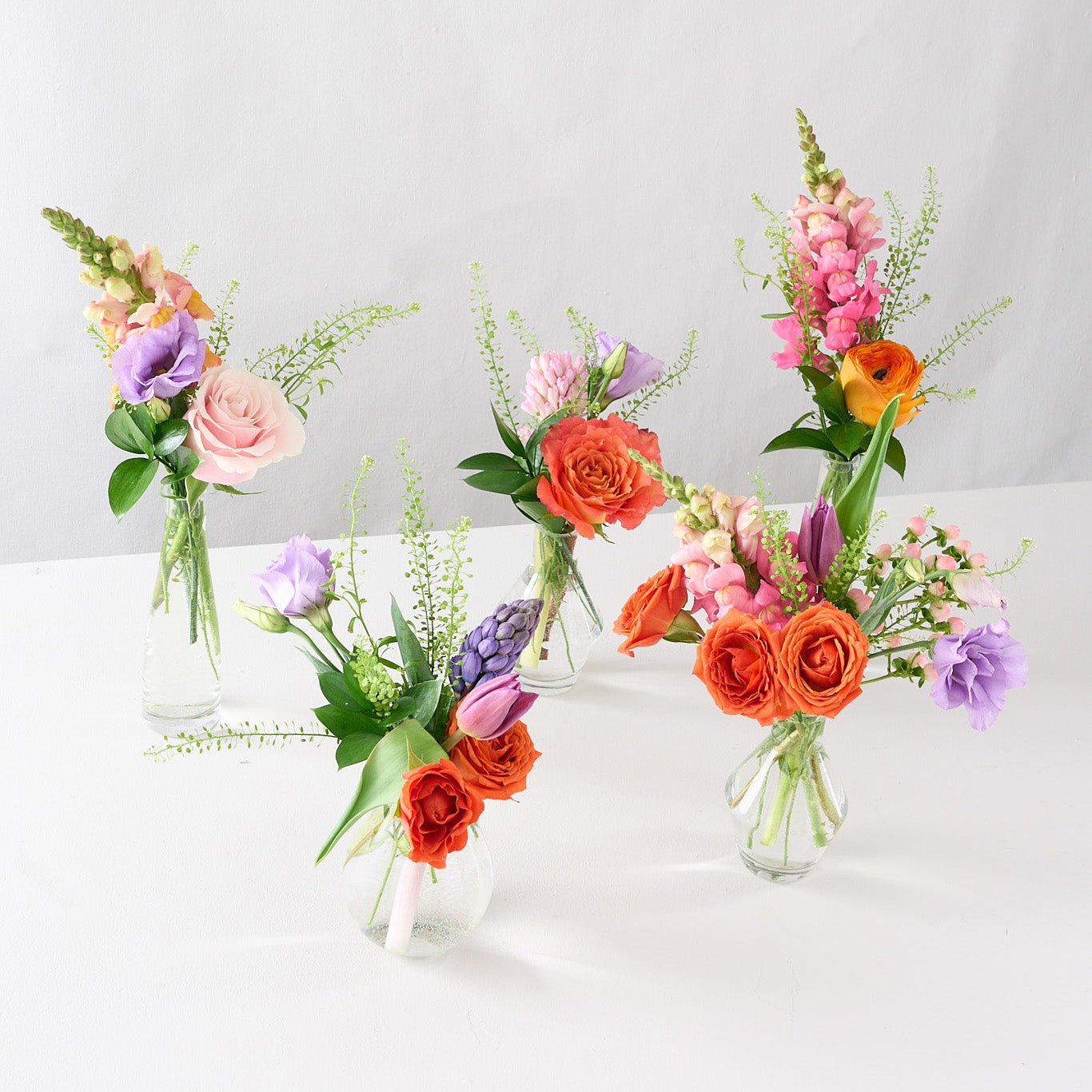 A series of five glass bud vases, each containing an arrangement of colorful spring flowers, arranged in a line on a light background.