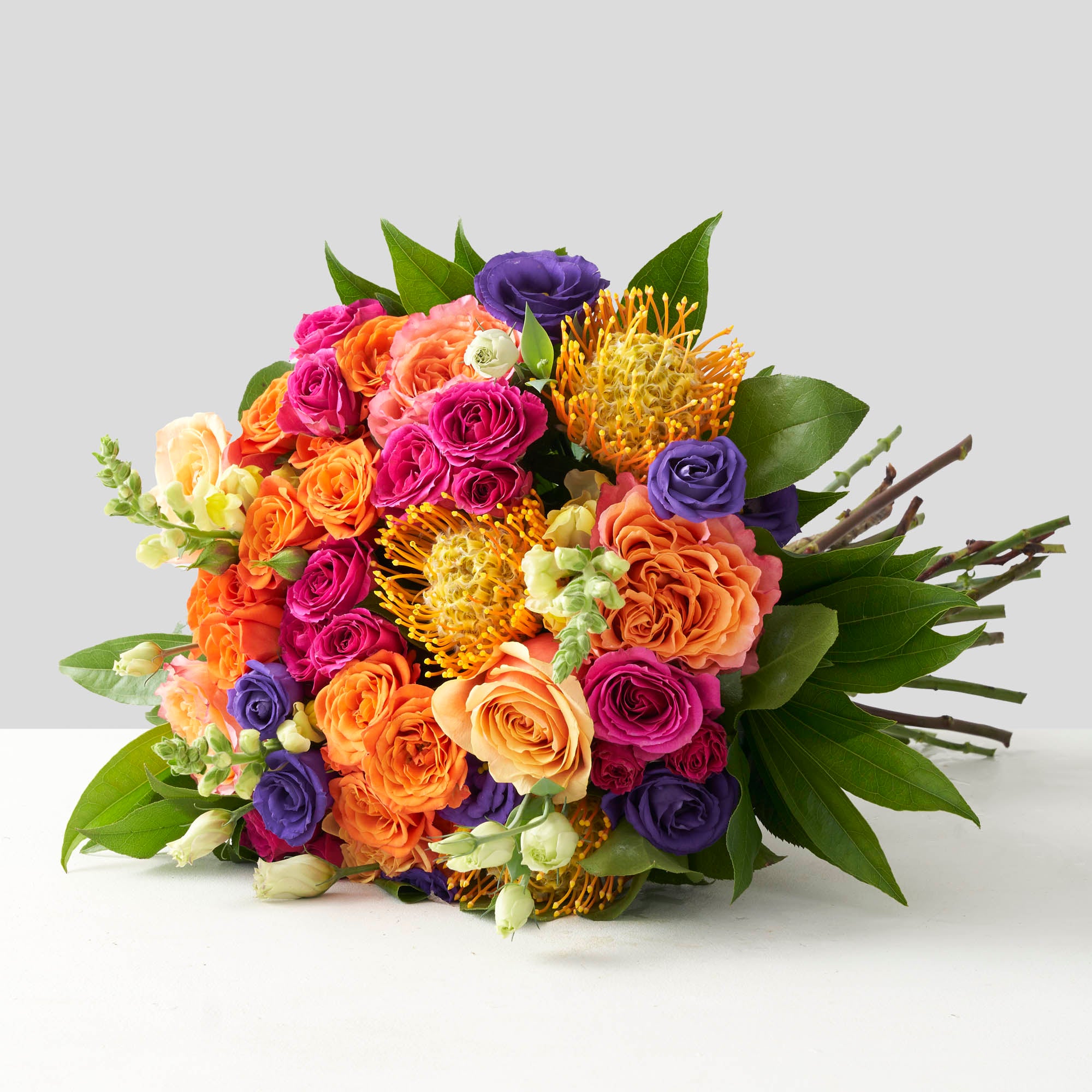 Round handtied bouquet with orange free spirit roses, dark purple lisianthus, hot pink spray roses, and gold protea.