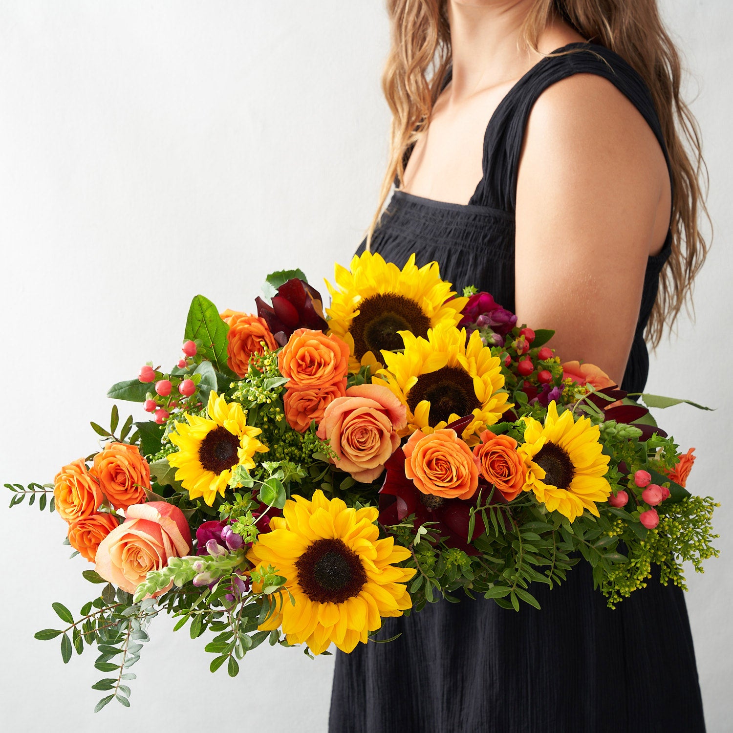 Woman in black dress holding a bouquet with sunflowers, free spirit roses, kahala roses, red hypericum berries, violet anemones, and other greenery