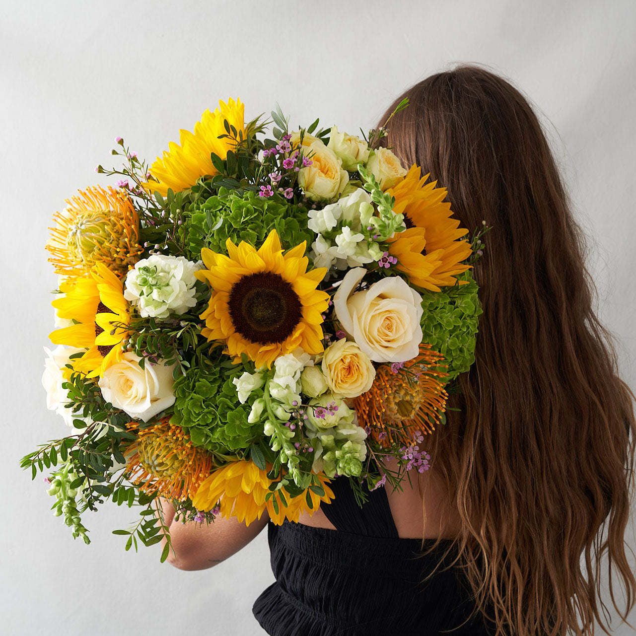 Woman in black top holding a bouquet of bright sunflowers, hydrangeas, white roses, spray roses, proteas, and other greenery