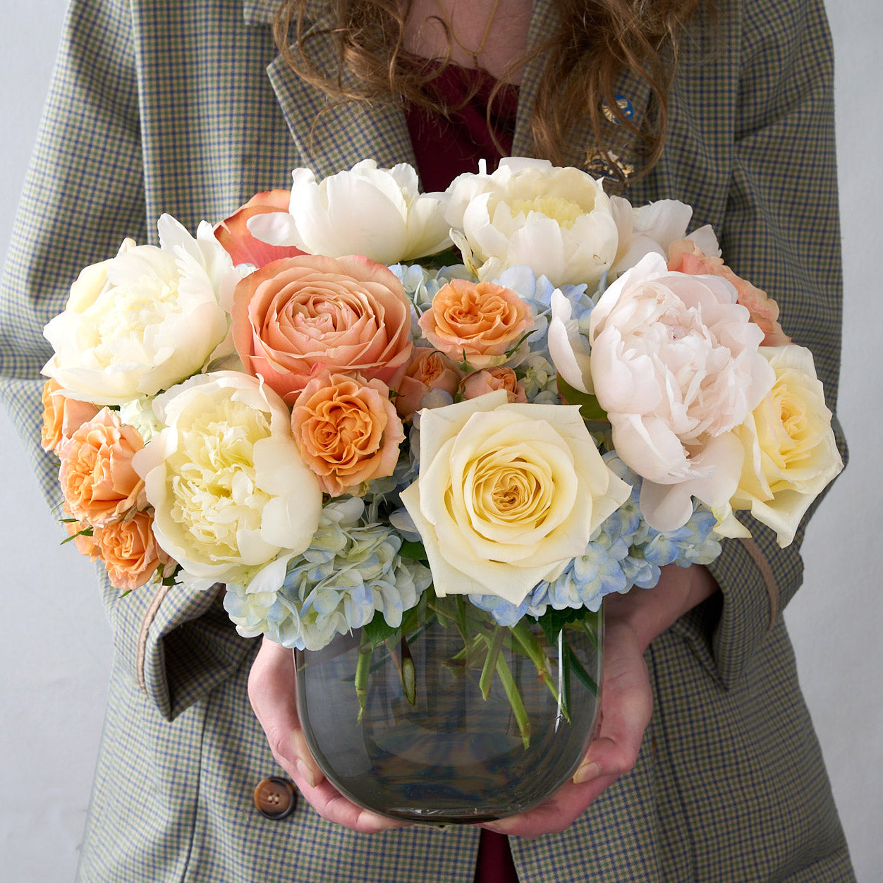 Woman in coat holding a glass vase that includes white and peach roses, cream peonies, and light blue hydrangeas