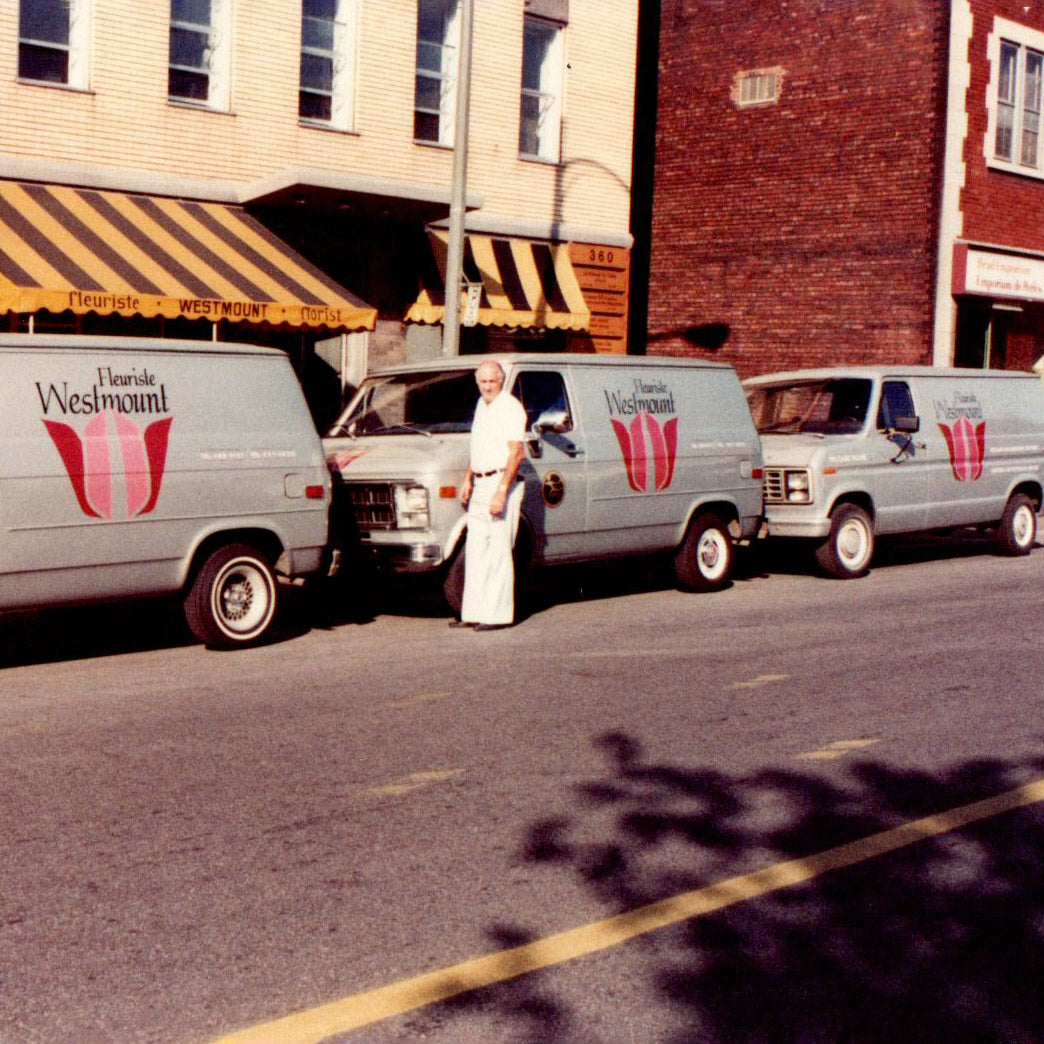 Old photo of man in wide legged pants in front of three grey vans with Westmount florist logo on them