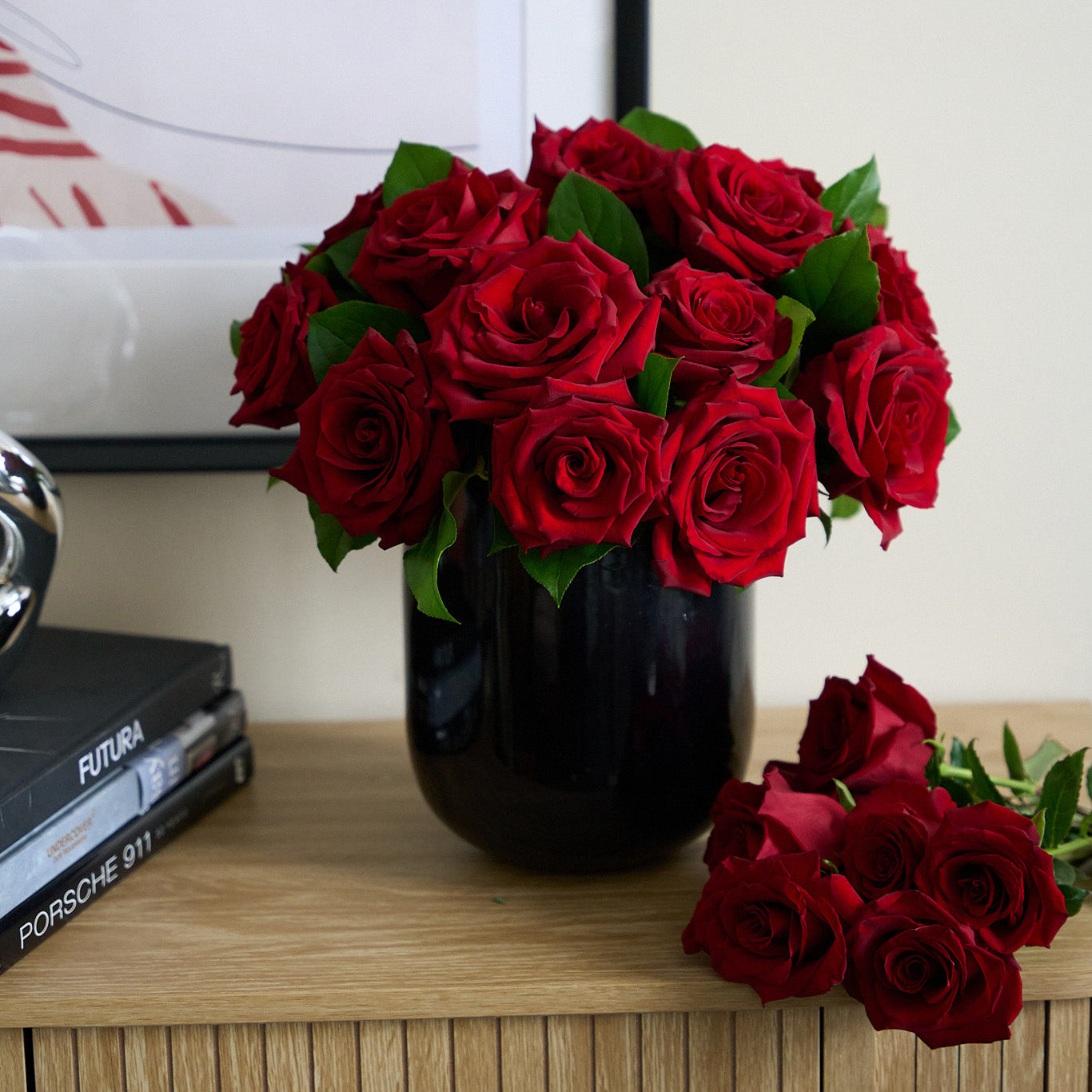 arrangement of red  valentine's day roses in a black glass vase on a credenza with 6 cut red roses laying next to it.