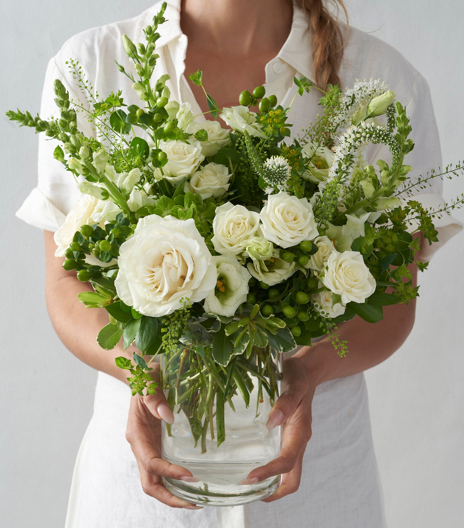 Woman wearing all white, holding clear glass vase filled with all white and green flowers including white roses, snapdragons, and lisianthus. 