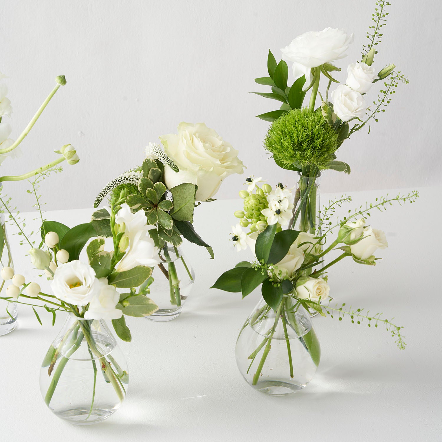 Four small clear glass vases filled with white and green flowers on white background.
