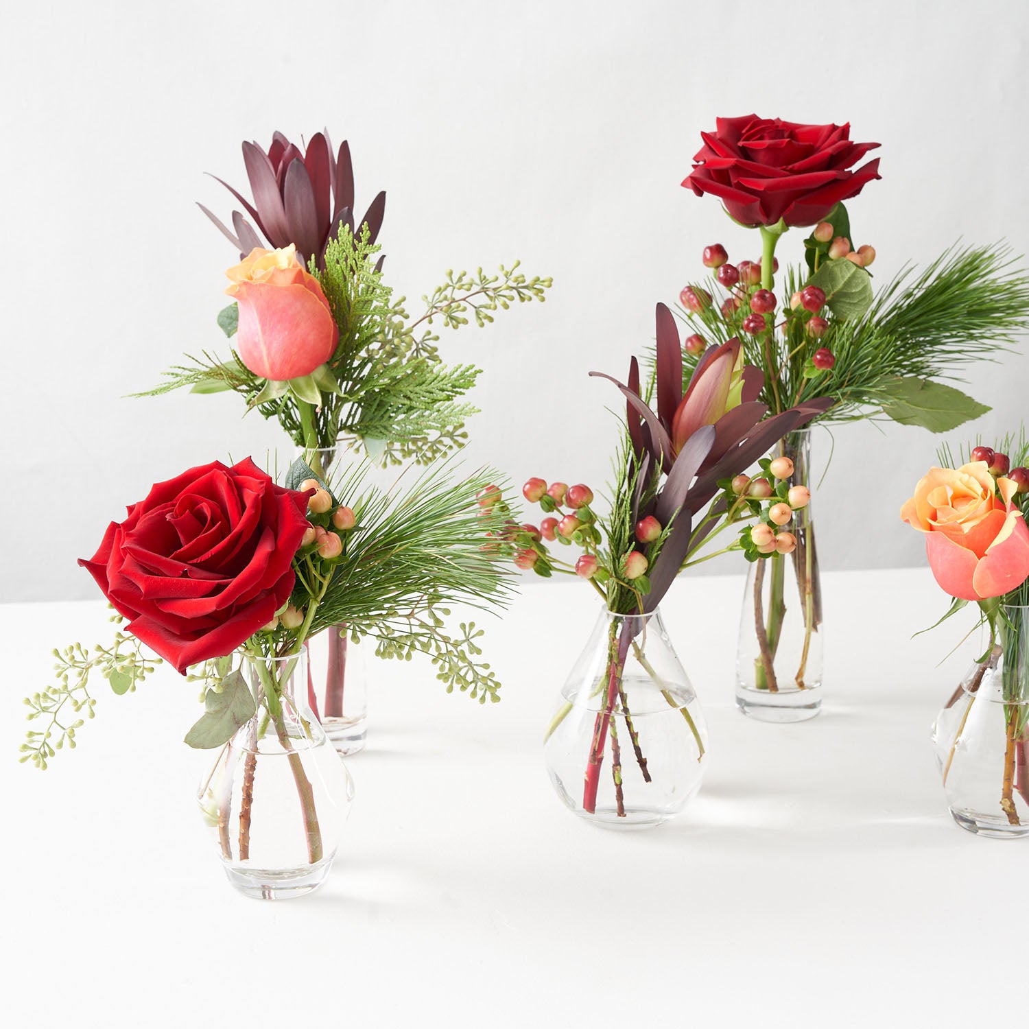 Five small glass vases filled with red roses, pine, Cherrybrandy roses, and hypericum berries.